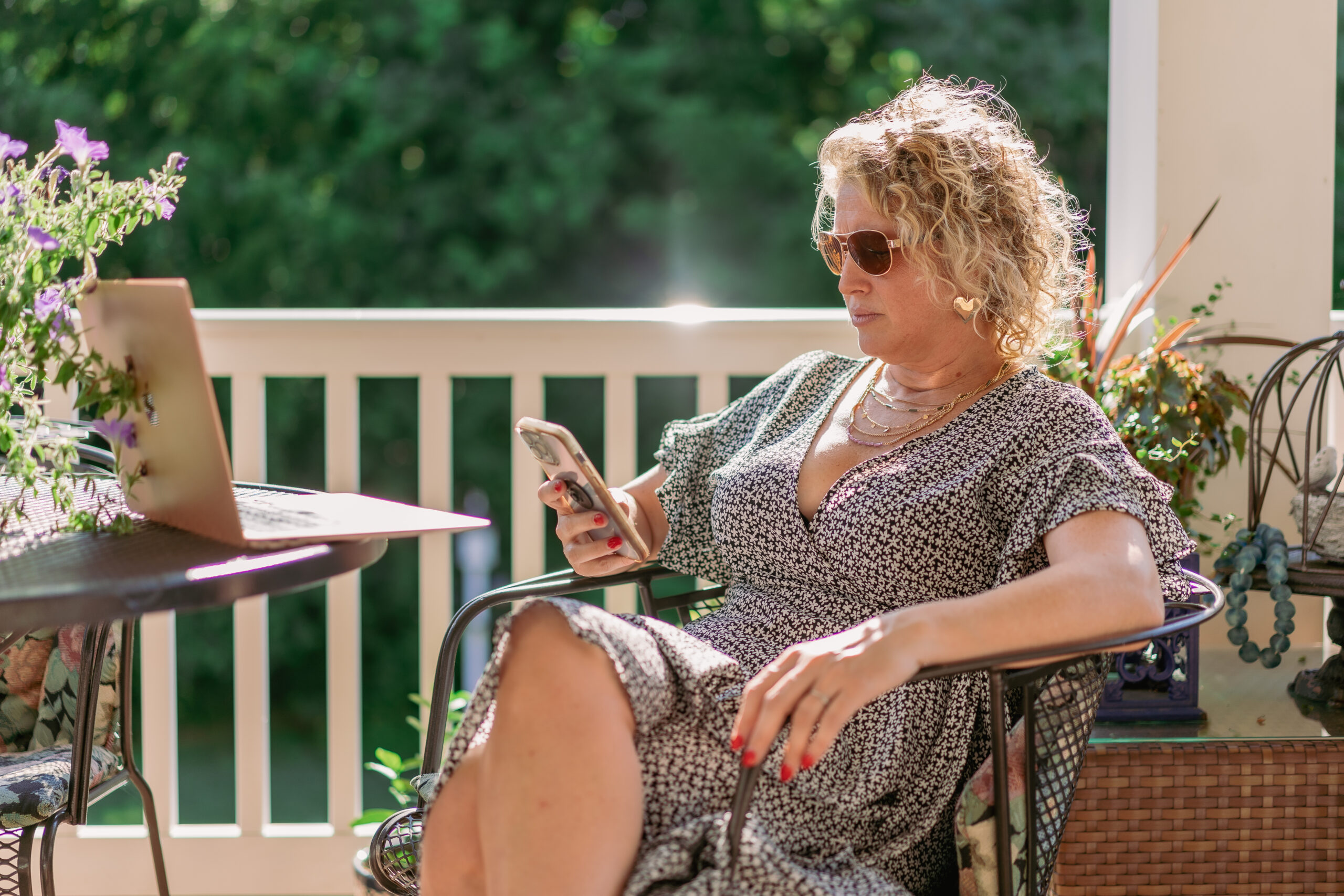 personal branding photo for realtors showing a female realtor sitting on her porch looking at her phone wearing a flowered dress and aviator sunglasses