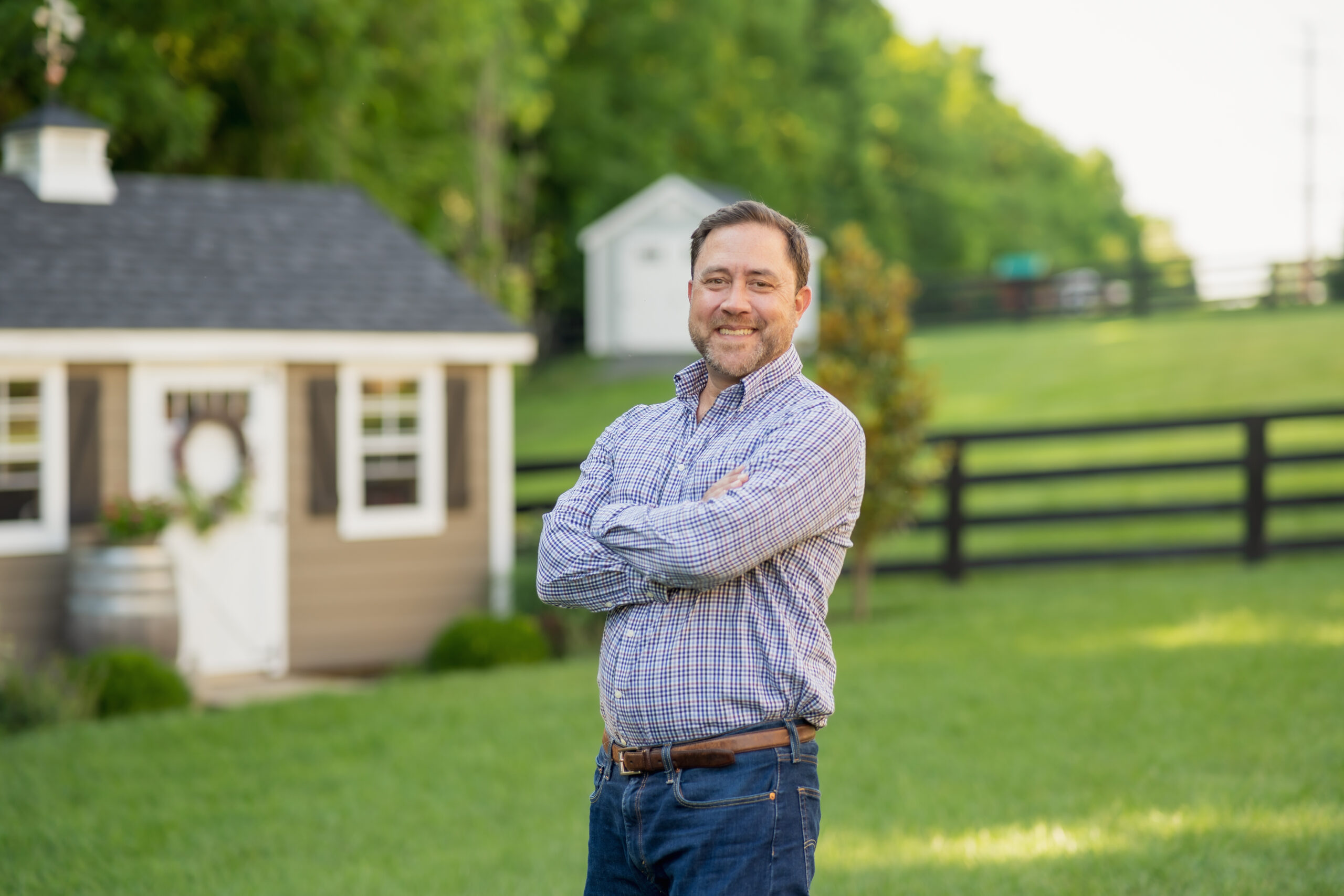 personal branding photo for realtors showing smiling male realtor in striped shirt with his arms crossed in a country setting with fenced fields in the background