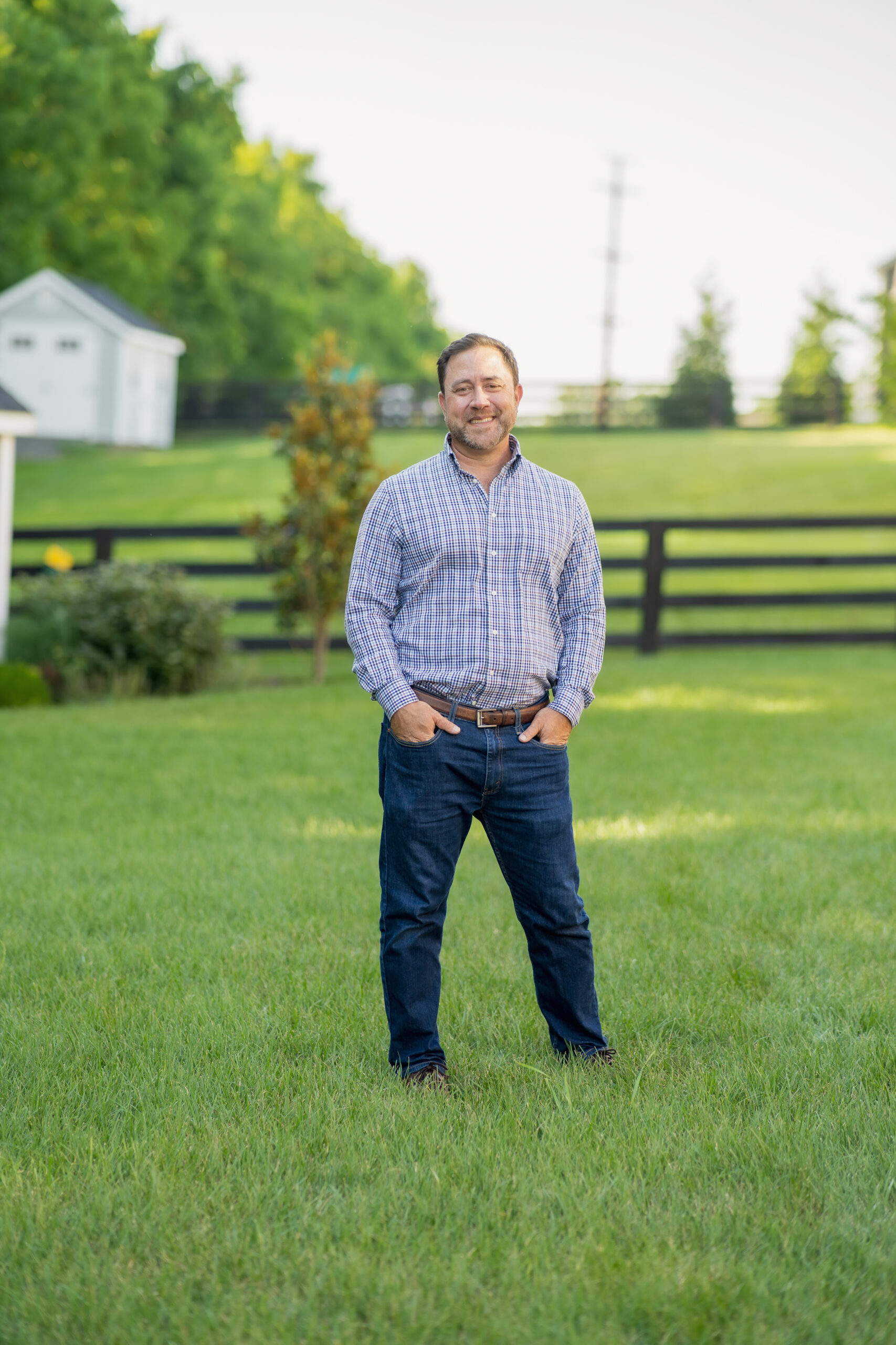 personal branding photo for realtors showing smiling male realtor in striped shirt with his hands in his pockets in a country setting with fenced fields in the background