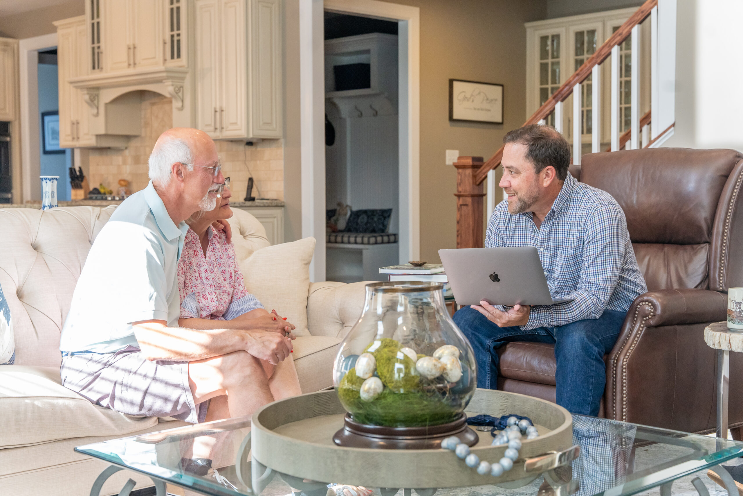 personal branding photo for realtors showing a smiling male realtor with his MacBook laptop sitting in the living room with 2 elderly clients