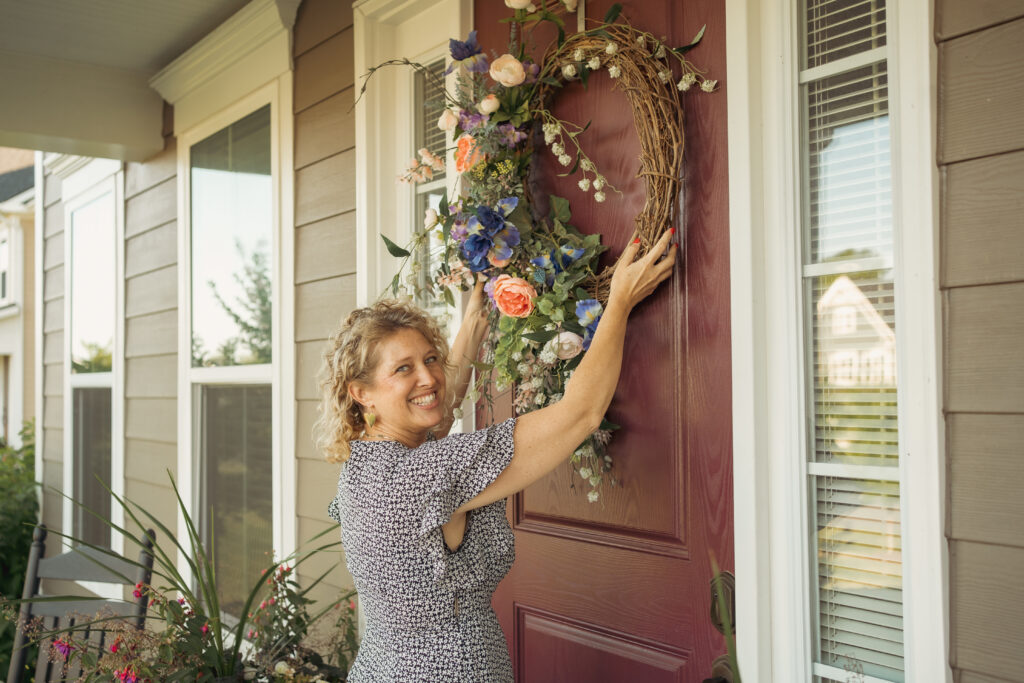 personal branding photo for realtors showing smiling female realtor in a flowered dress hanging a floral and wicker wreath on a burgundy colored front door