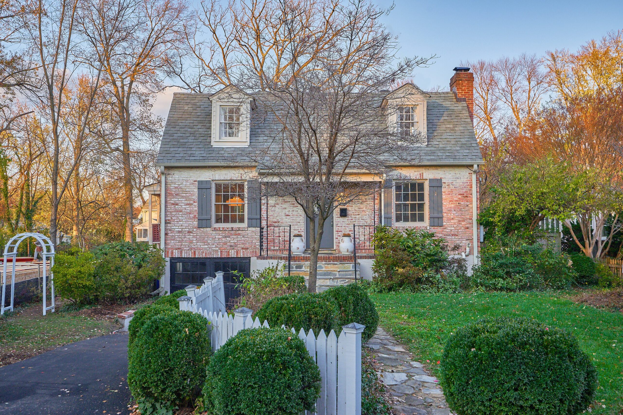 Professional exterior photo of 5933 16th St N, Arlington, VA - showing the front. Home is a white washed red brick Cape Cod with grey shingle roof and stone front steps. A stone pathway leads from the road straight to the steps.