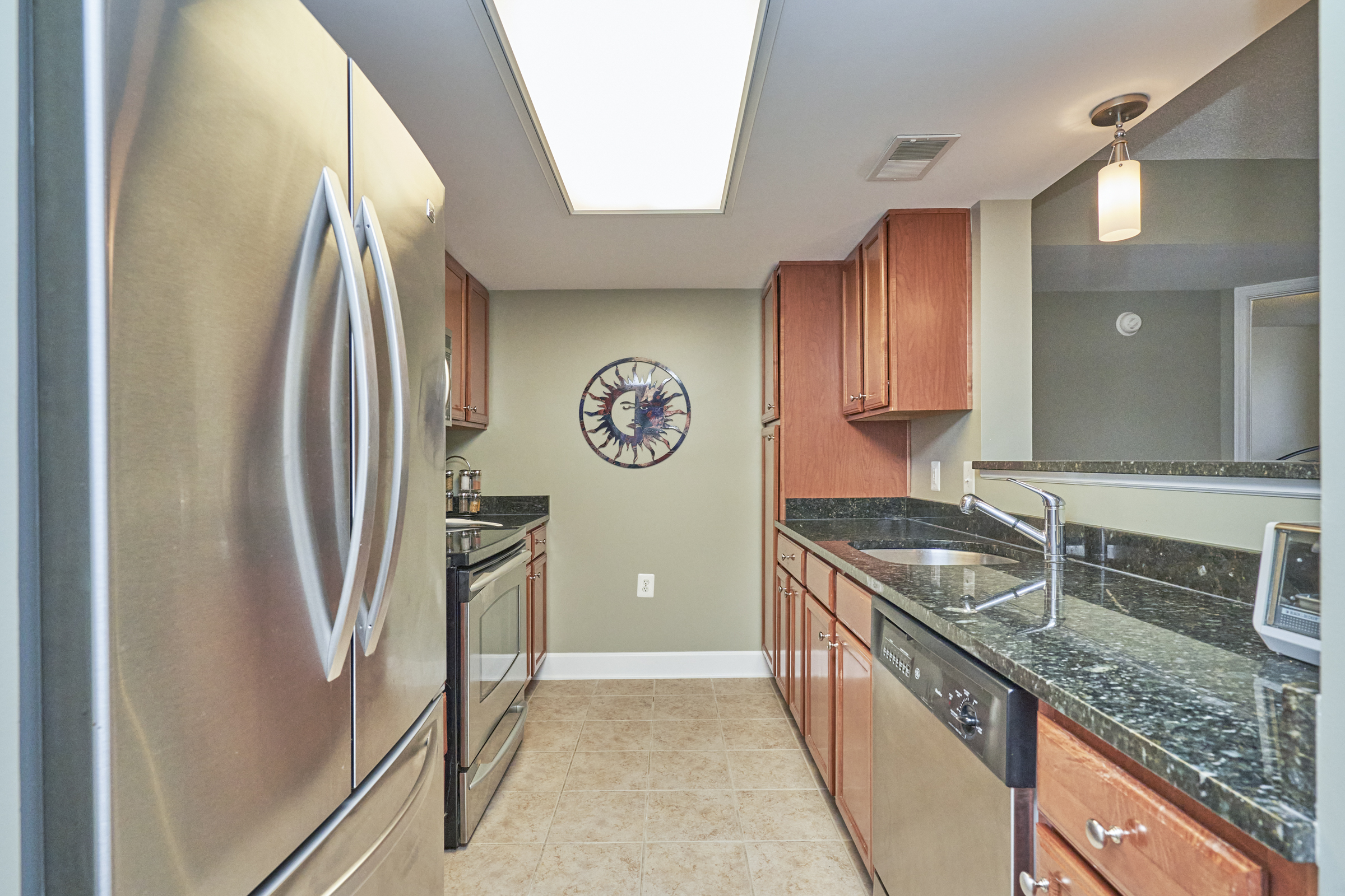 Interior professional photo of 11800 Sunset Hills Road Unit #811, showing the kitchen from the perspective of looking in from the hall. Stainless appliances and dark granite counters are seen with walnut cabinets