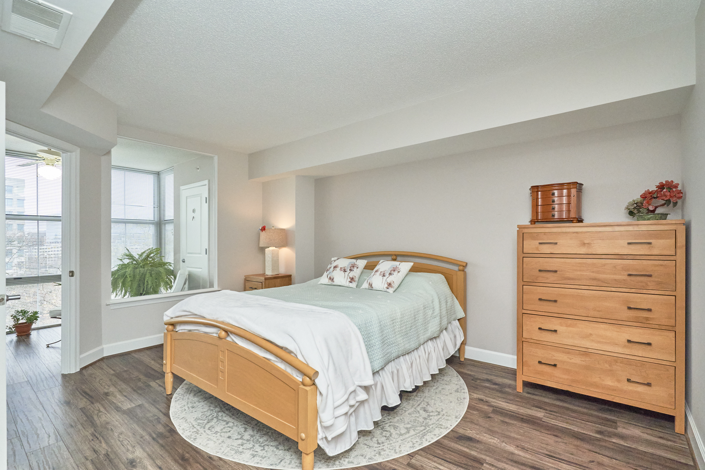 Interior professional photo of 11800 Sunset Hills Road Unit #811, showing one of the bedrooms with recessed ceiling, bed and dresser in the foreground, and view of the connected sunroom in the background/left angle