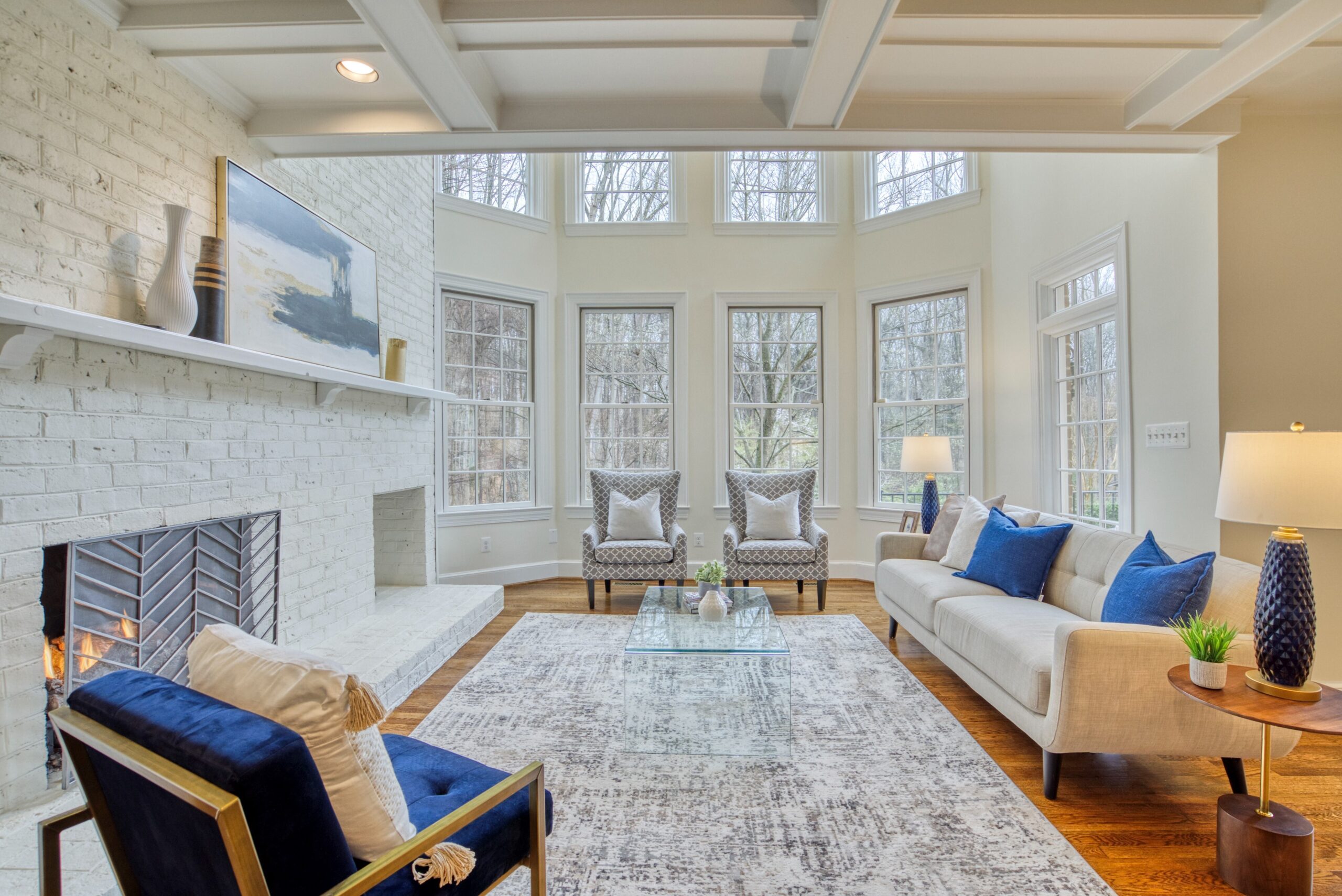 Professional interior photo of 718 Potomac Knolls Dr - showing the great room with white brick fireplace, hardwood floors, coffered ceiling and soaring 2-story windows and tasteful cream and navy furniture.