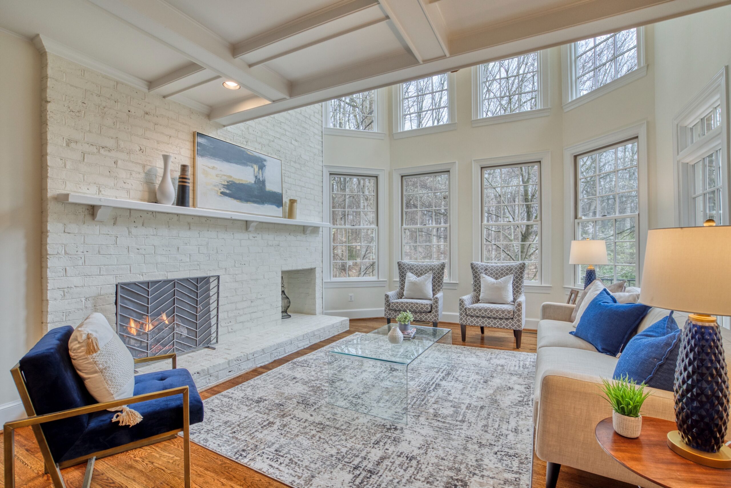 Professional interior photo of 718 Potomac Knolls Dr - showing the great room with white brick fireplace, hardwood floors, coffered ceiling and soaring 2-story windows and tasteful cream and navy furniture.