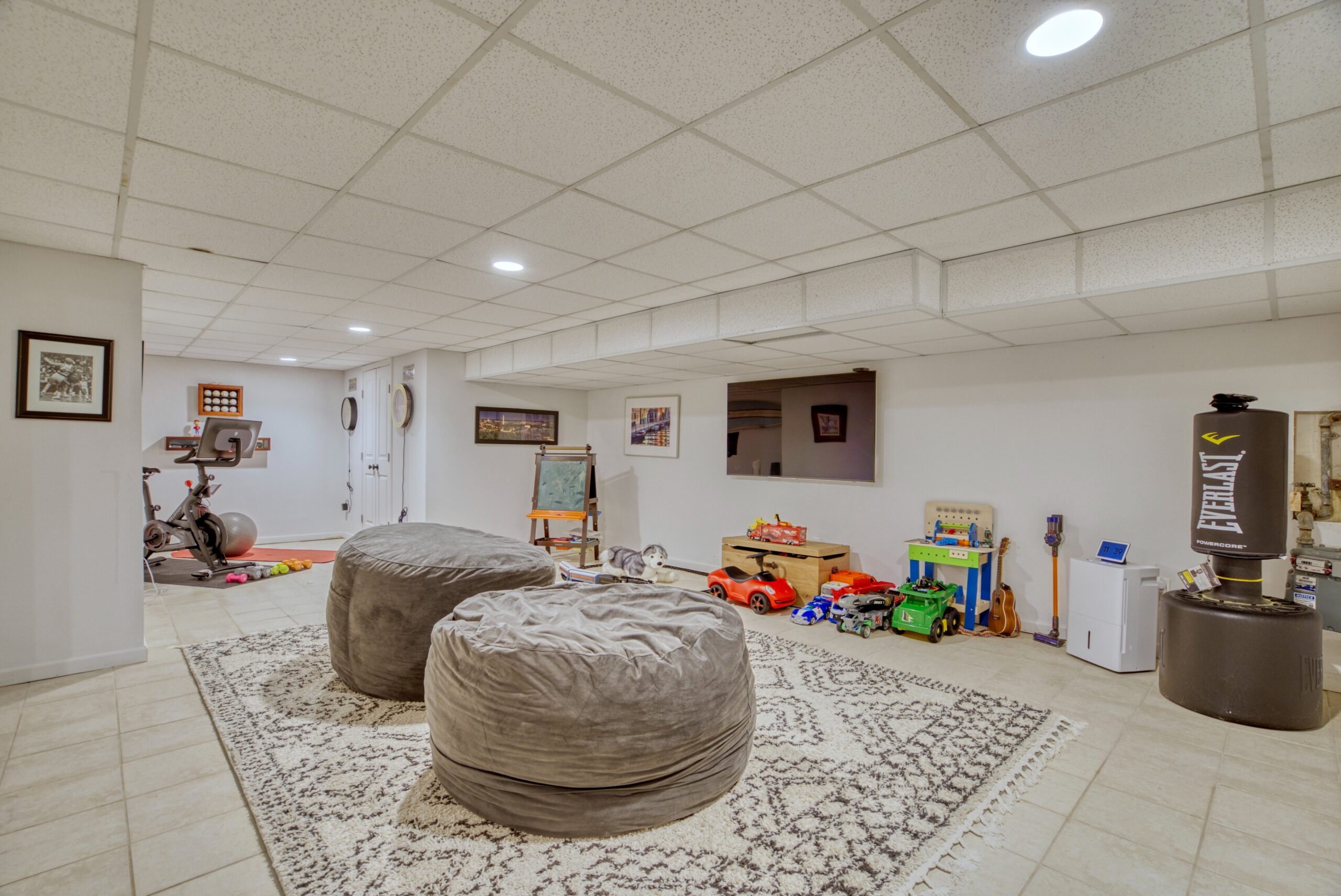 Professional interior photo of 209 Norwood Road, Annapolis, MD - showing the basement rec-room with tile floors, ceiling tiles, area rug and various children's toys and workout equipment