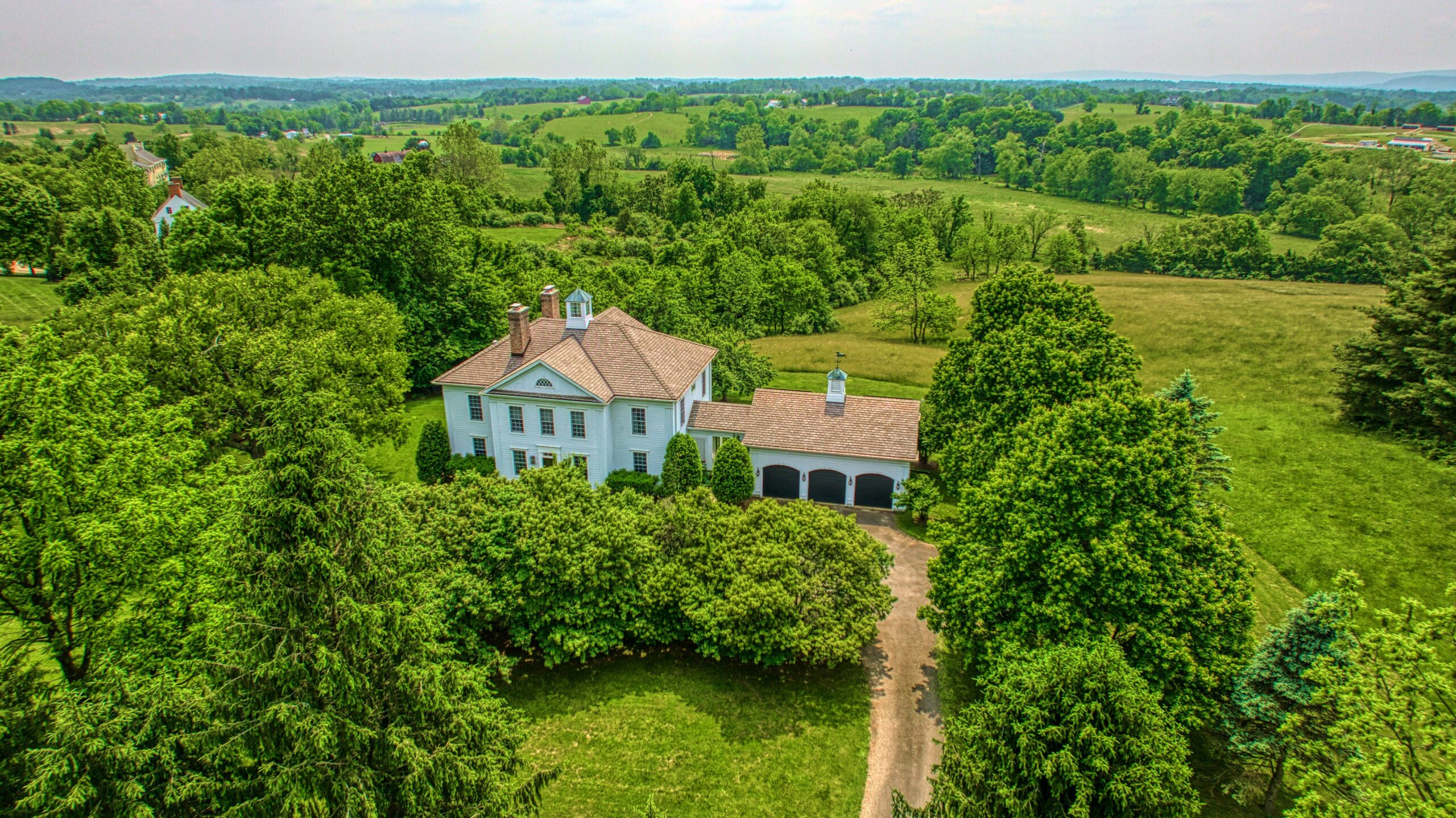 Professional drone aerial photo of 15203 Clover Hill Road - showing the front of the white home surrounded by trees and 3-bay garage visible