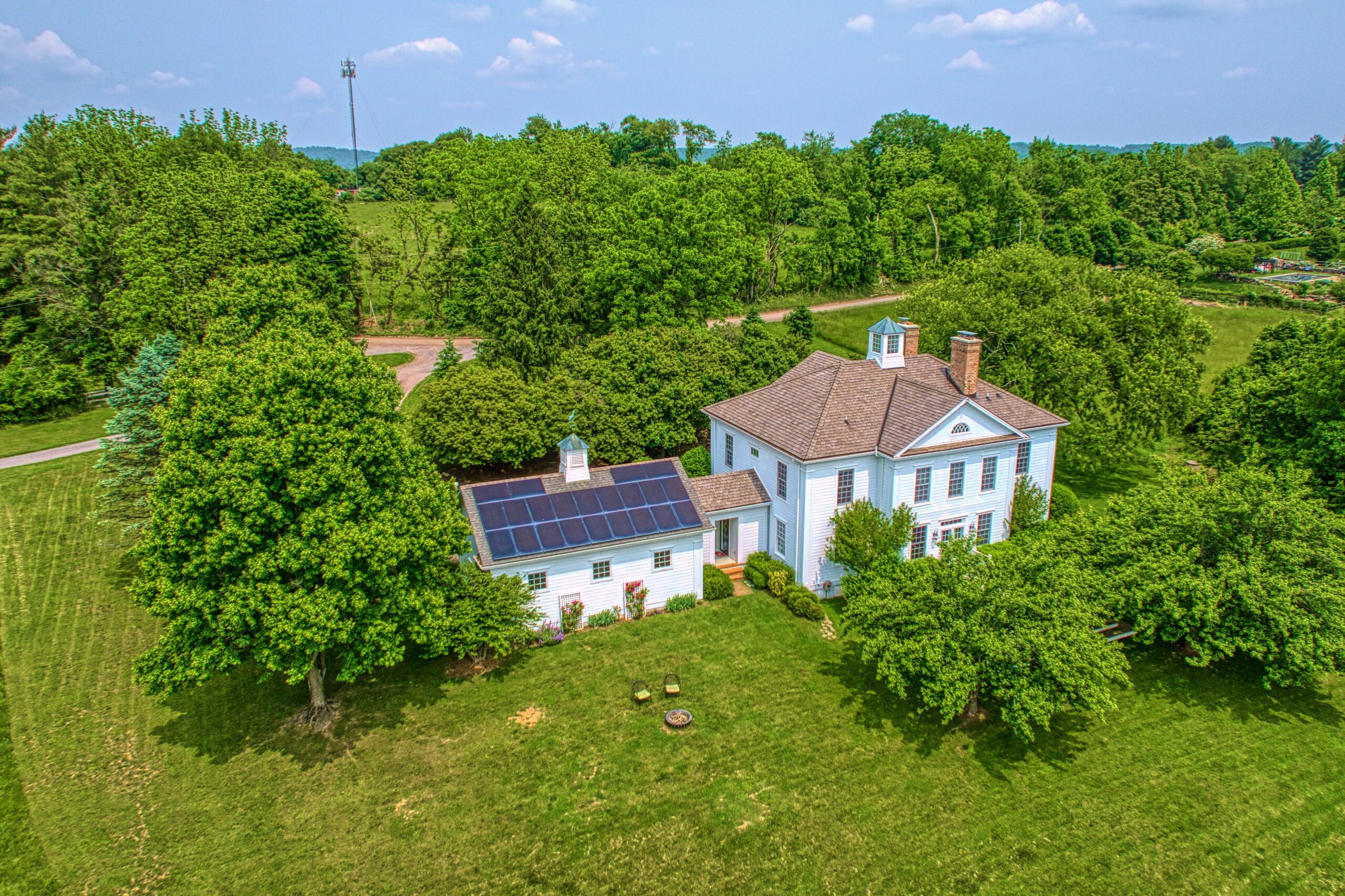Professional drone aerial photo of 15203 Clover Hill Road - showing the rear of the white home surrounded by trees and 3-bay garage visible with solar panels