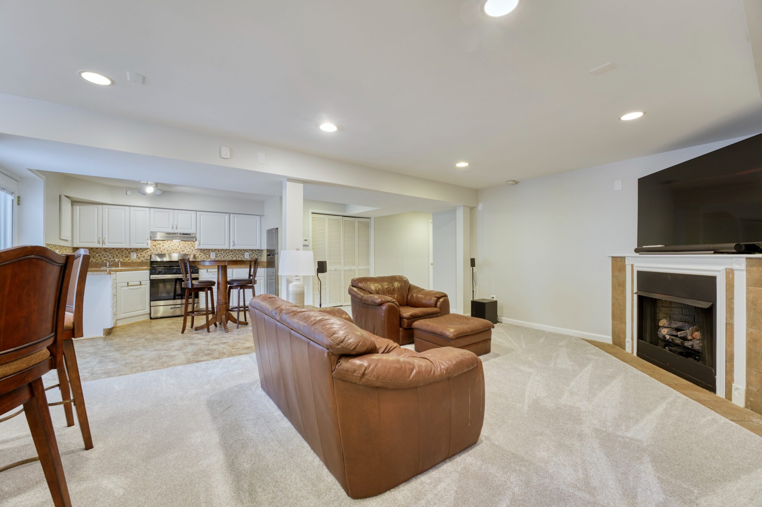 Professional interior photo of 13700 Holly Forest Dr - showing the basement with a carpeted living room with fireplace in the foreground and a second kitchen in the background on tile floors. 