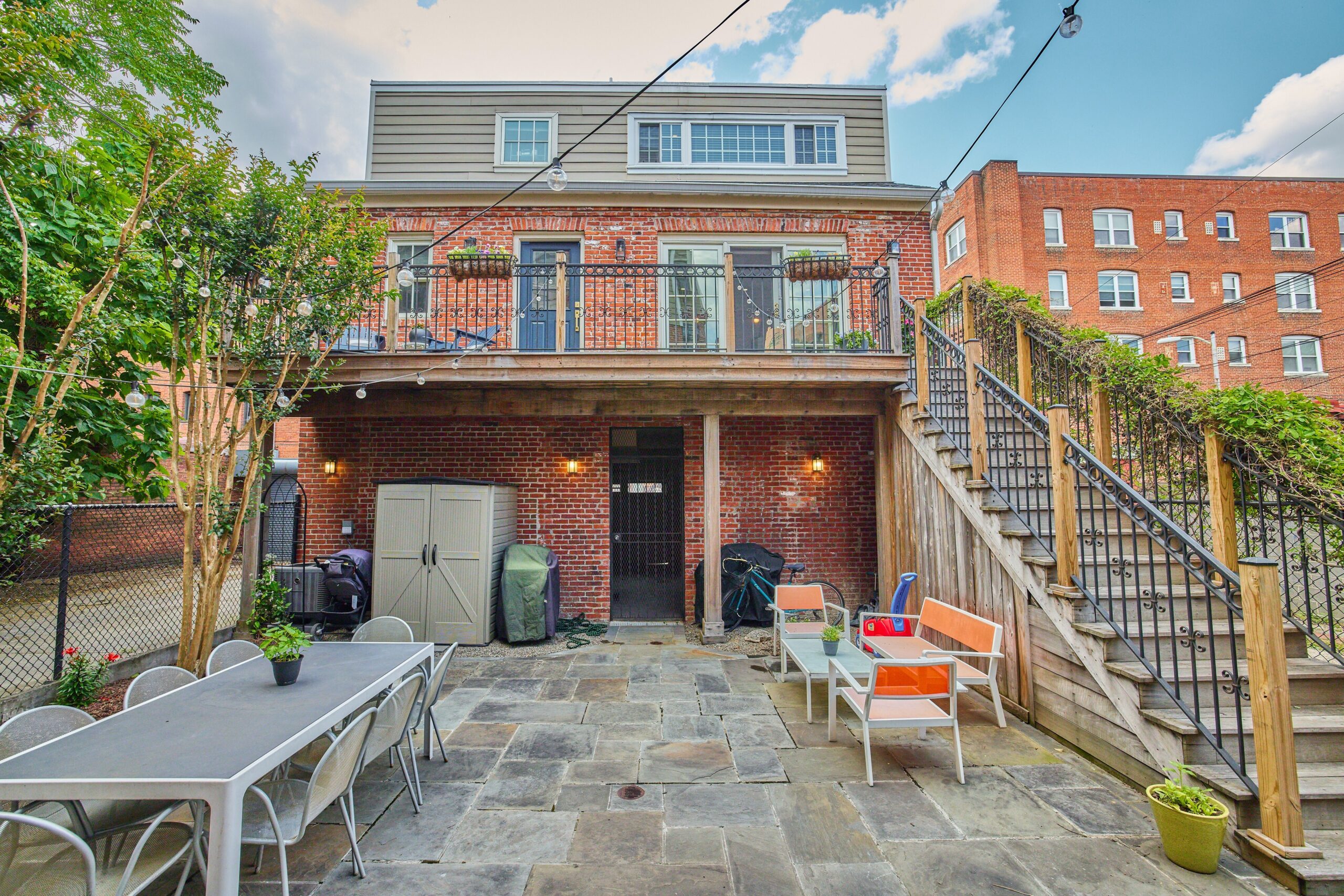 Professional exterior photo of 1459 Harvard St NW #6 - showing the rear of the building where the entrance to #6 is. Expansive stone patio with storage area, and staircase up to balcony where the entrance is.