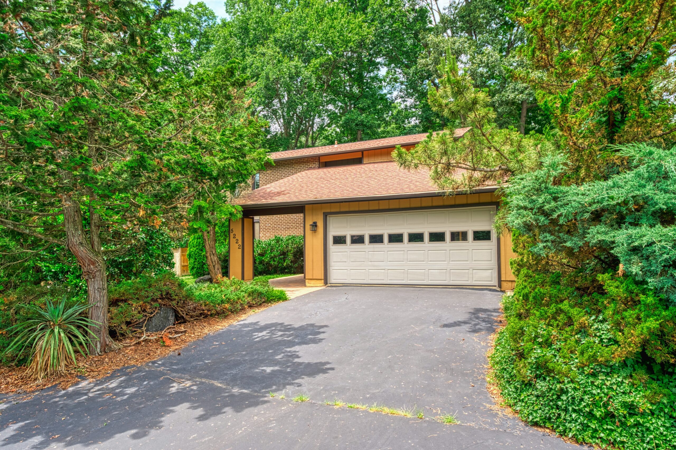 Professional exterior photo of 5222 Bradfield Drive in Burke, Virginia - showing the street view with mature trees that surround the contemporary home. The 2-car garage is visible along with the entrance to the covered walkway to the entrance