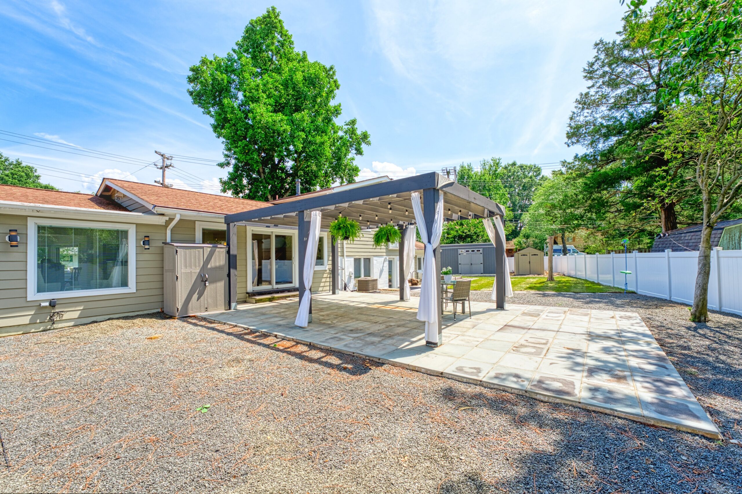 Professional exterior photo of 8320 Fort Hunt Rd, Alexandria - showing the backyard from one corner of the fenced lot with a large pergola on an even larger patio with lush landscaping in the background