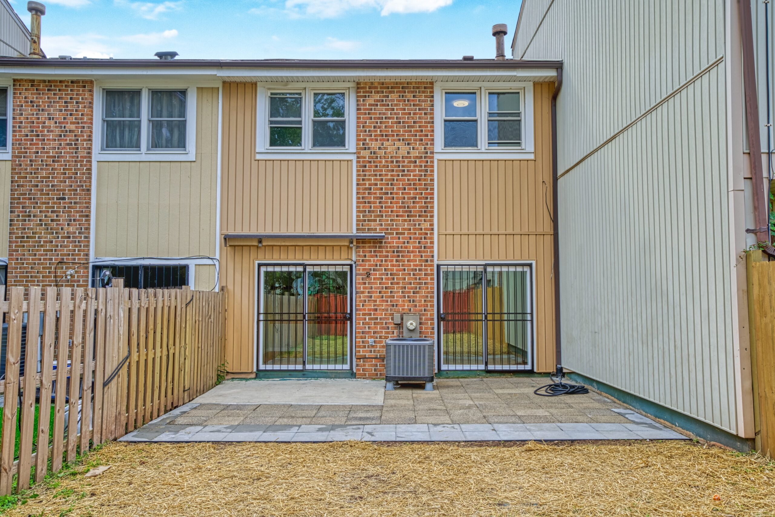Professional exterior photo of 6302 Arwen Court, Fort Washington, MD - showing the rear of a townhome with tan vertical siding and brown brick with 2 double sliding door exits to a patio
