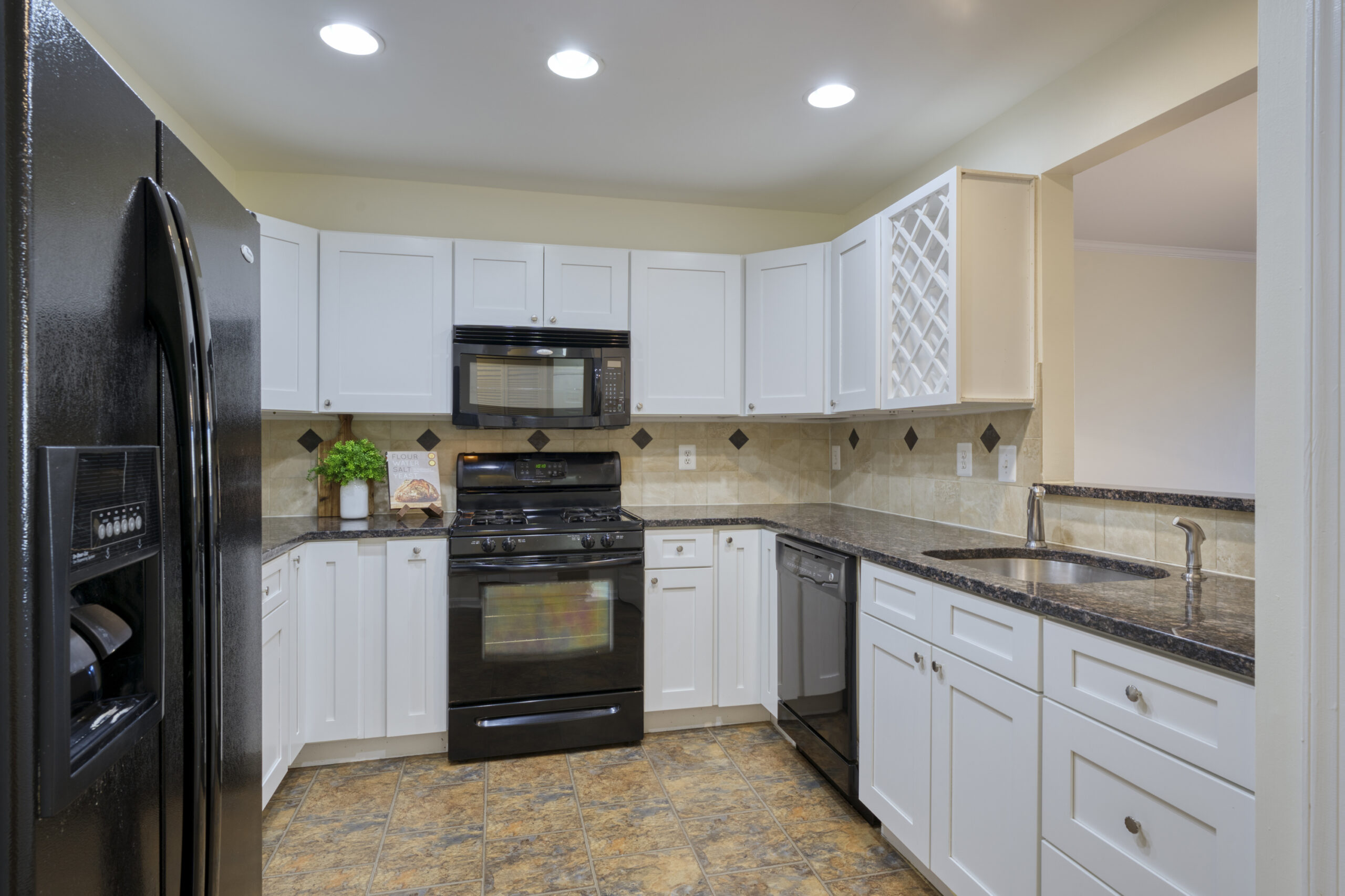 Professional interior photo of 20588 Cornstalk Ter Unit 101 - Showing the kitchen with black appliances, white cabinets, a picture window to the living room and black granite counters