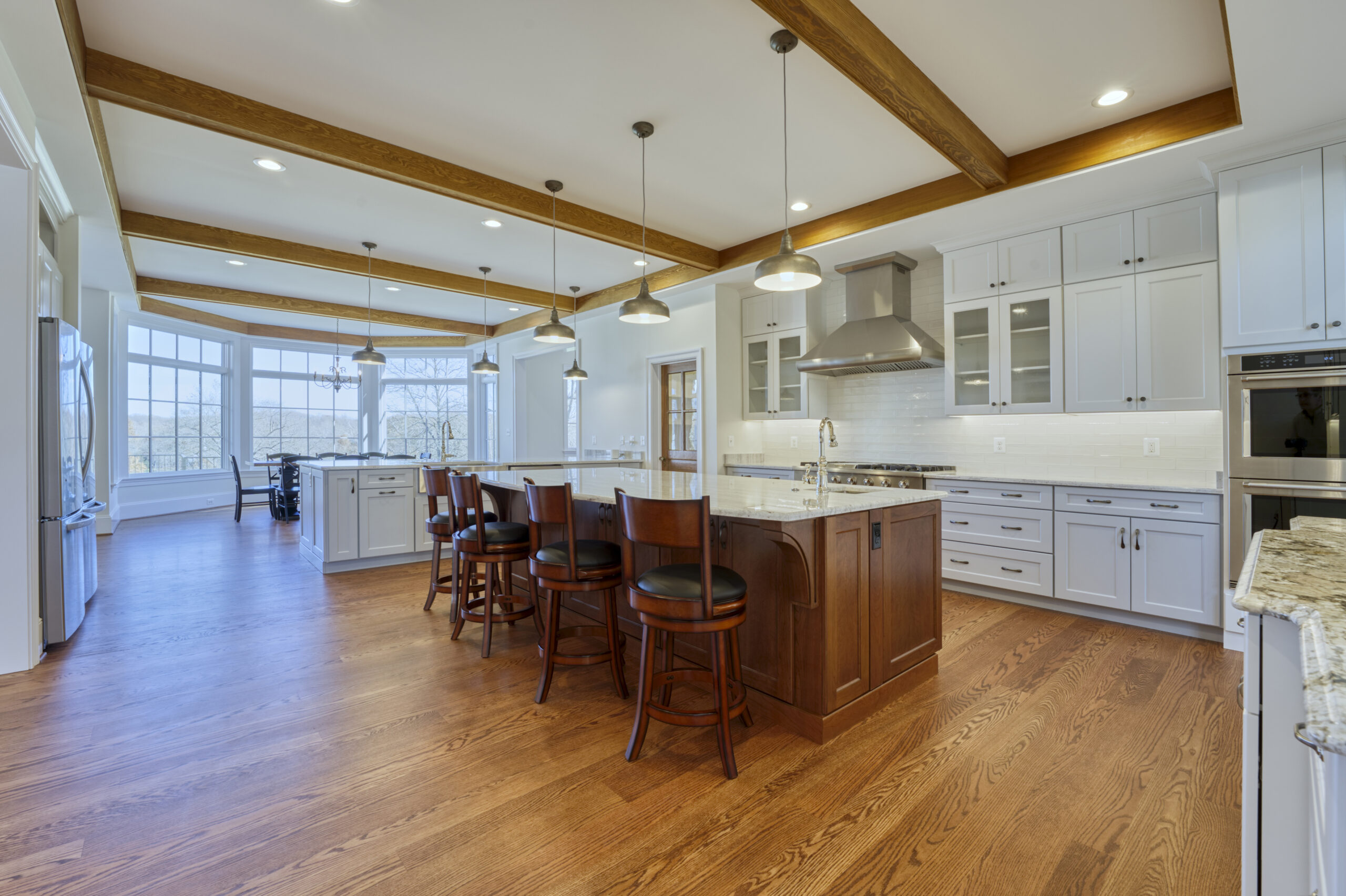 Professional interior photo of custom build new home by Black Oak Construction - showing the kitchen with exposed wood beams in the white ceiling, large island centered in front of the range with stainless hood, wrap around counters with stainless appliances and farmer's sink and hardwood floors
