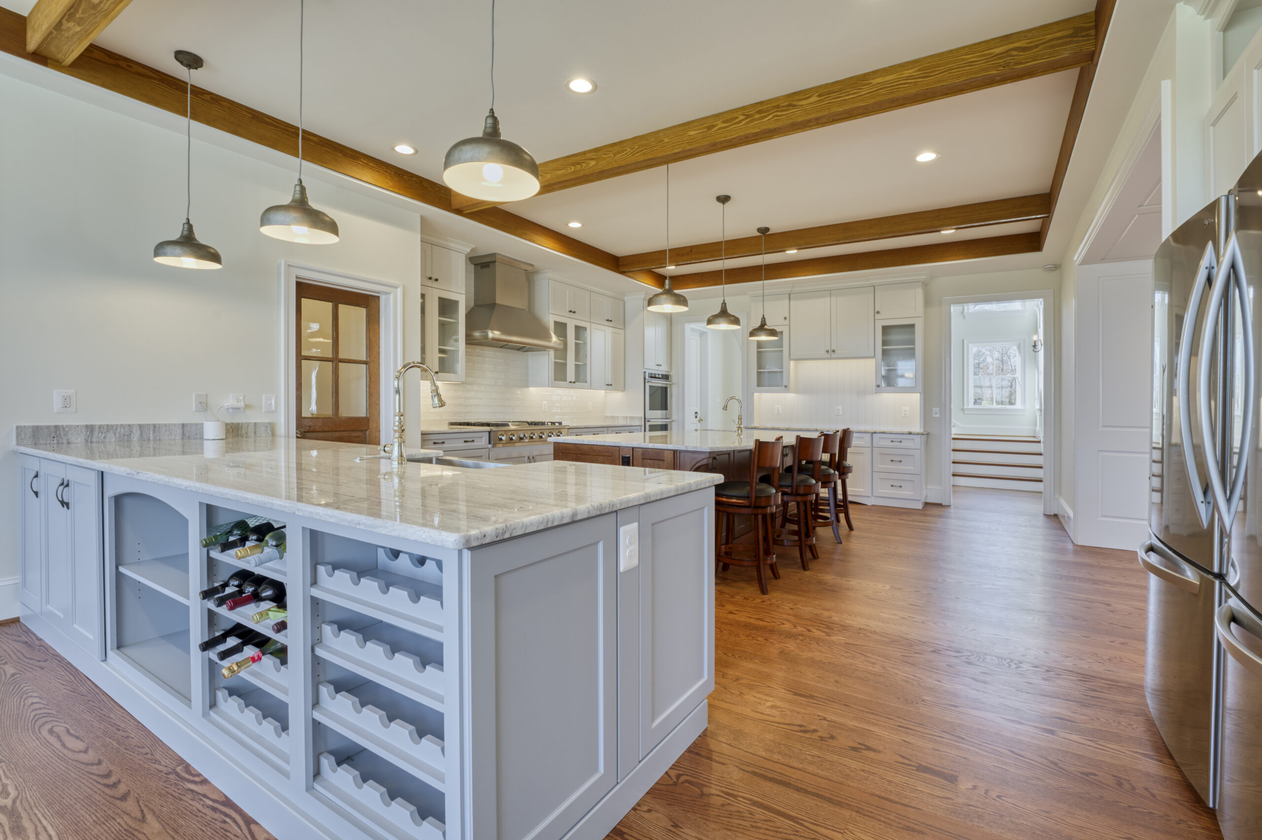 Professional interior photo of custom build new home by Black Oak Construction - showing the kitchen with exposed wood beams in the white ceiling, large island centered in front of the range with stainless hood, wrap around counters with stainless appliances and farmer's sink and hardwood floors. built in wine racks in the foreground